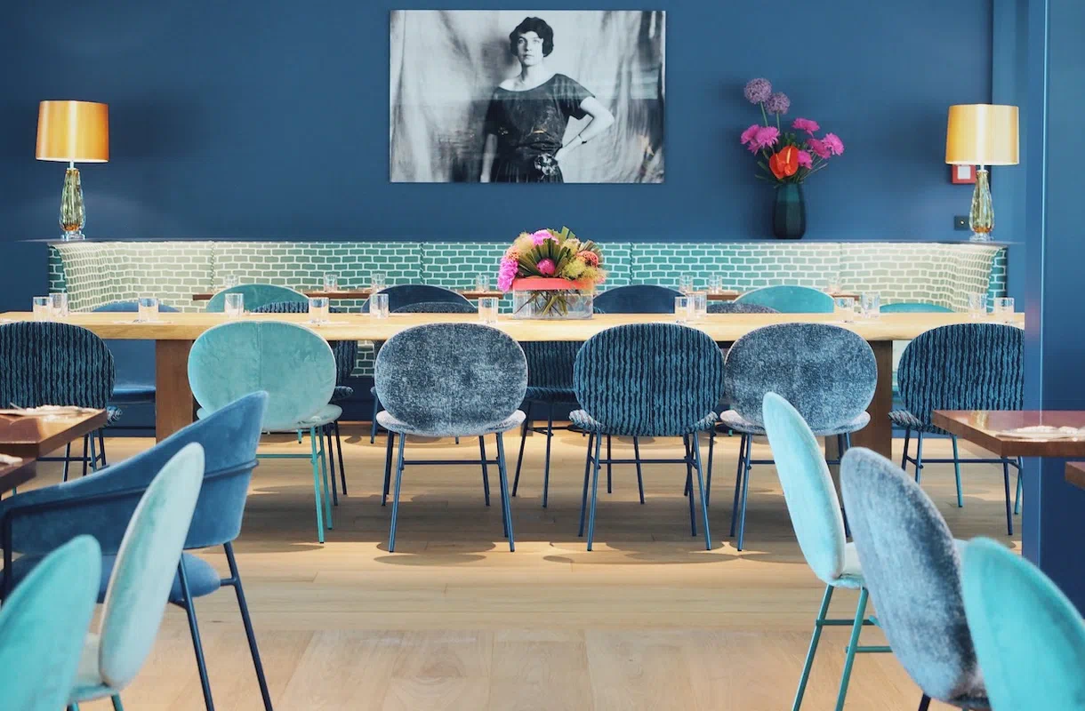 Geräumiger Frühstücksraum mit blauen Samtstühlen, einem langen Holztisch mit Blumenarrangement im Augustin Hotel