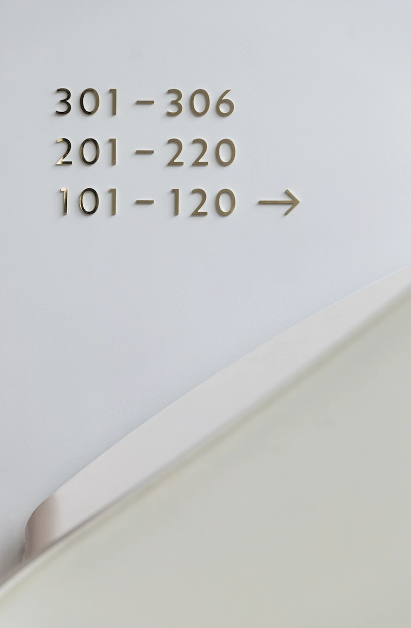 Minimalistischer Zimmernummern-Wegweiser im Augustin Hotel, mit goldenen Zahlen auf weißer Wand und modernem Design.