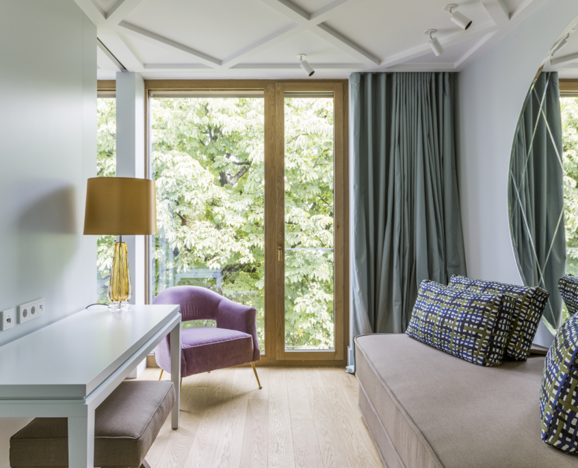 lichtdurchflutetes Wohnzimmer mit bodentiefen Fenstern und Blick ins Grüne, ausgestattet mit einem Schreibtisch, einem Sessel und einem gemütlichen Sofa